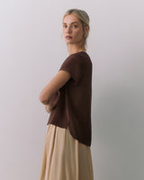 Person in a short sleeve top and long skirt, standing sideways with arms crossed.