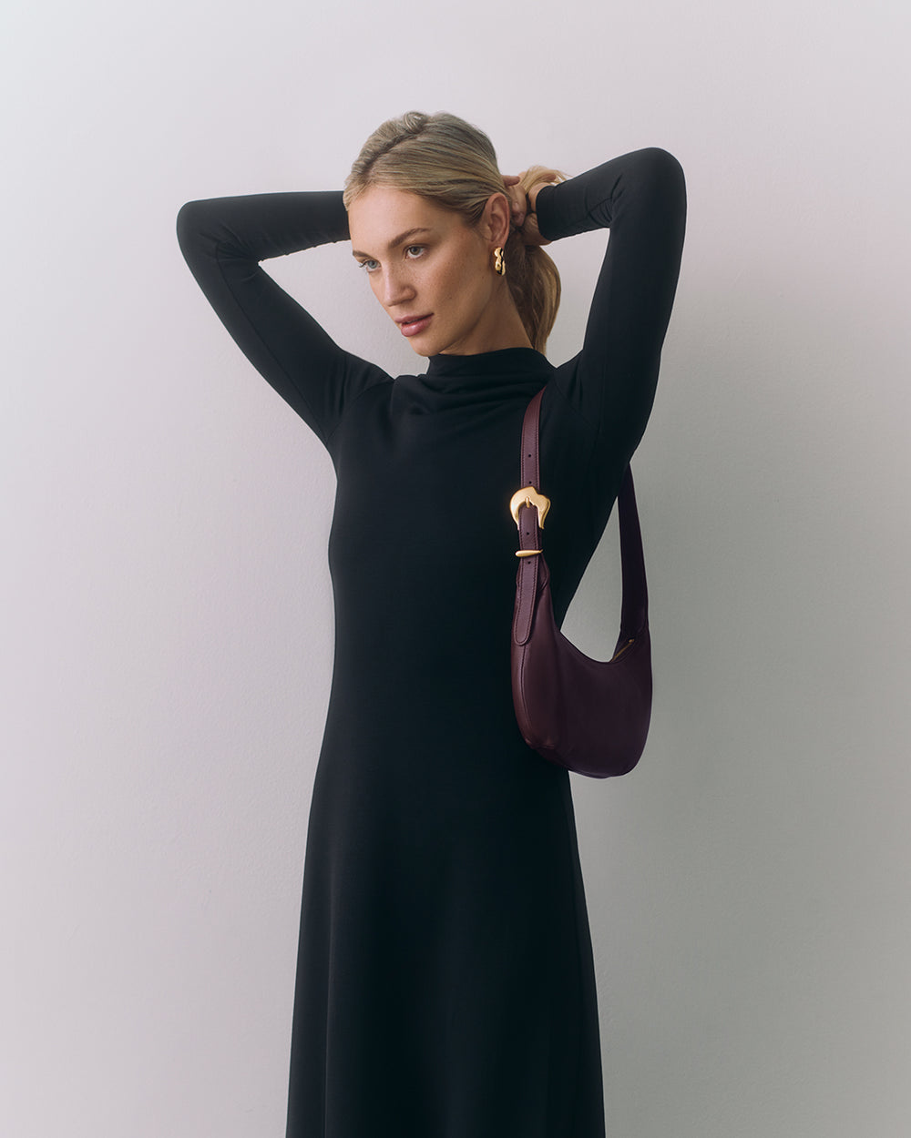 Person in long dress with small shoulder bag adjusts their hair with arms raised