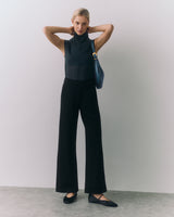 Woman standing with arms up, wearing sleeveless top and wide-leg pants, holding a shoulder bag.