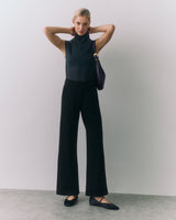 Person in sleeveless top and wide-leg pants, holding a shoulder bag, standing indoors.