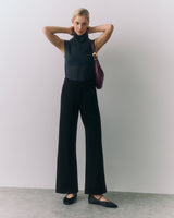 Woman stands with arms raised, wearing sleeveless top, pants, holding a shoulder bag.
