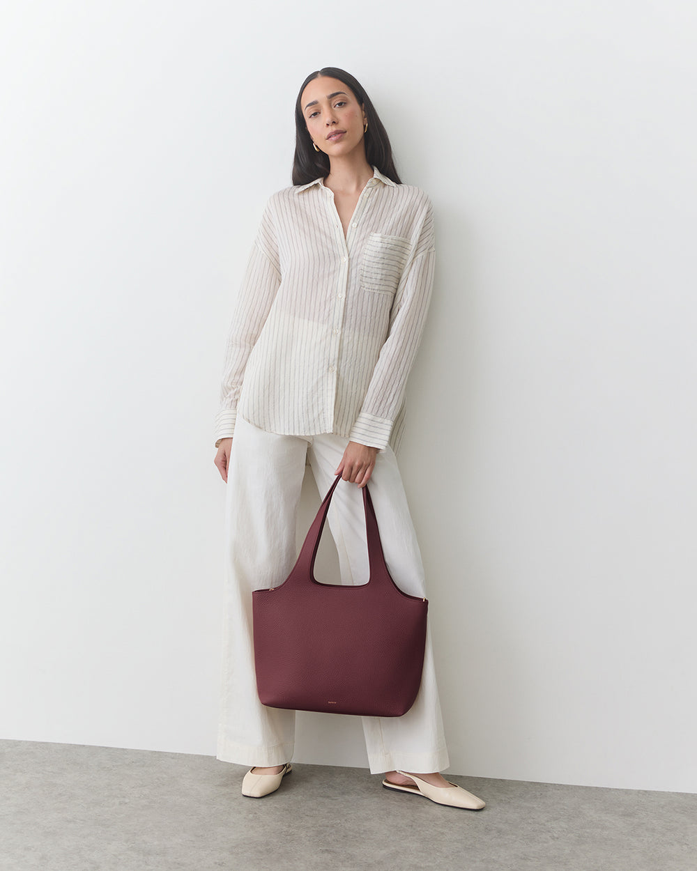 Person in striped shirt and wide pants holding a handbag, standing against a wall.