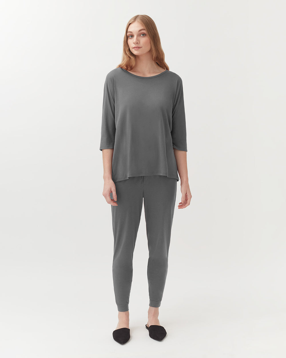 Woman standing in a casual outfit with long sleeves and fitted pants, wearing slippers.