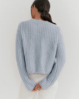 Woman standing with her back to the camera, wearing a sweater and with hands clasped behind her.