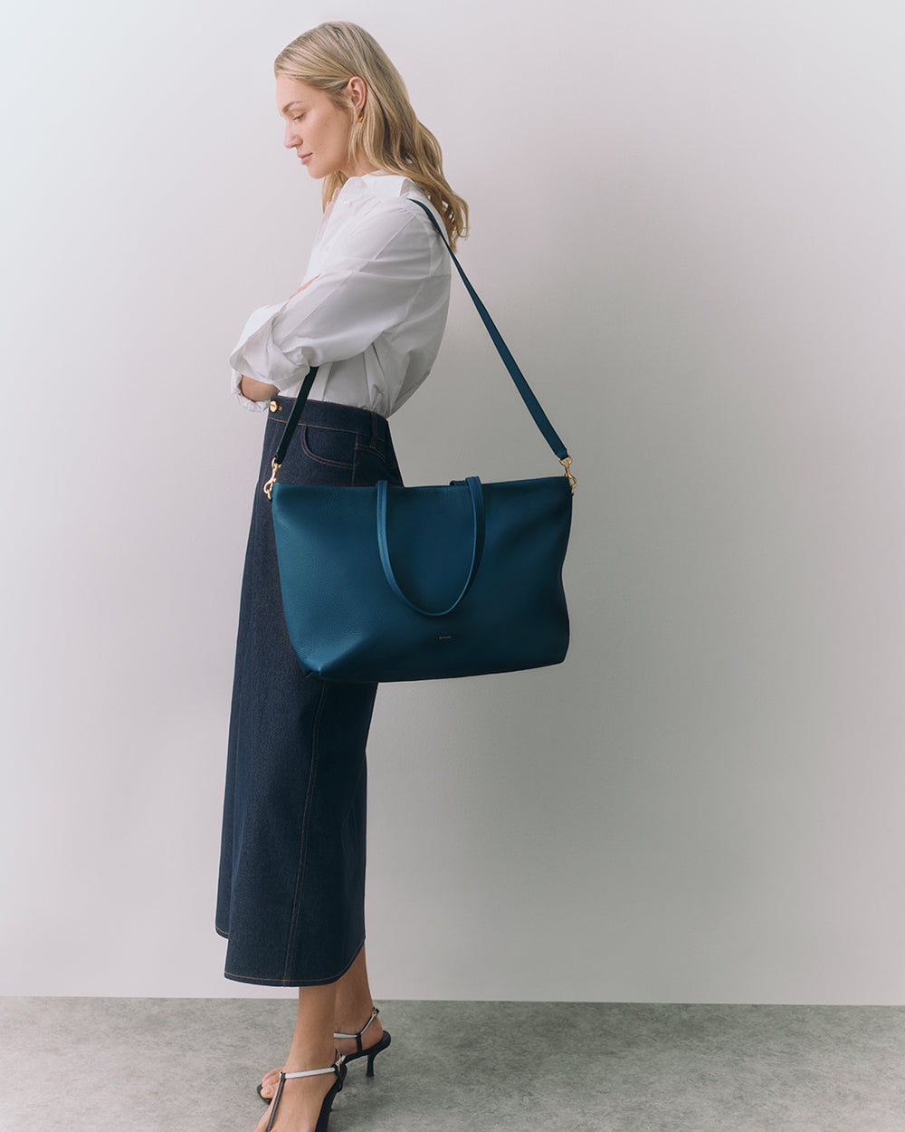 Person in long skirt holding large shoulder bag standing against plain background