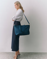 Person in long skirt holding large shoulder bag standing against plain background
