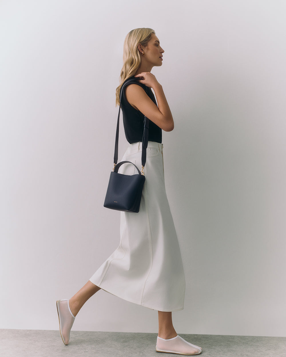 Person in sleeveless top and long skirt, holding a shoulder bag, walking indoors.