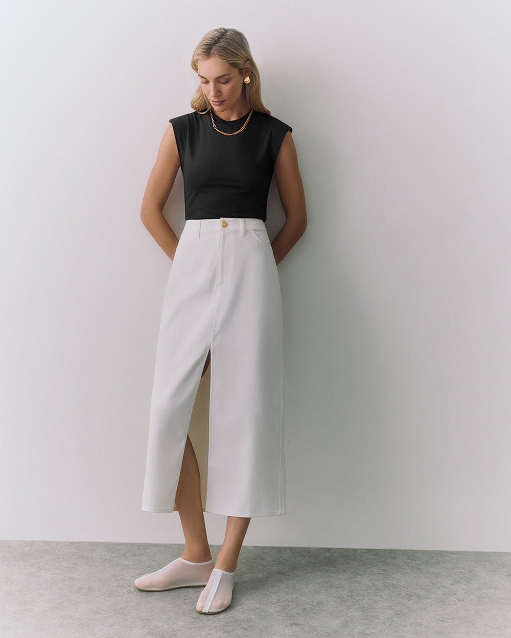 Person with sleeveless top and long skirt stands against a wall.