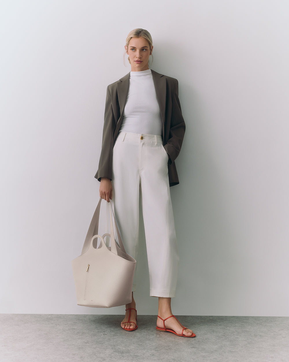 Person standing with a large handbag, wearing a blazer, top, pants, and open-toe shoes.