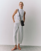 Person wearing sleeveless top, trousers, sandals, with crossbody bag, standing against plain backdrop.