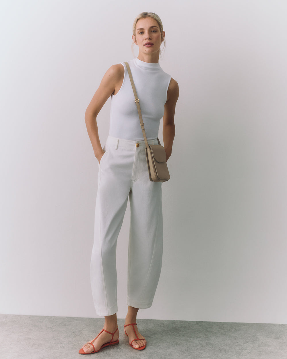 Person wearing sleeveless top, trousers, sandals, with crossbody bag, standing against plain backdrop.