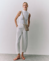 Person wearing sleeveless top, trousers, sandals, with crossbody bag, standing against plain backdrop.