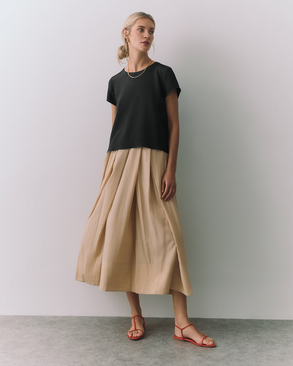 Person wearing a dark top with a long, flowing skirt and sandals, standing against a plain wall.