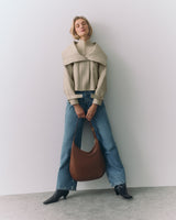 Person posing with a shoulder bag, wearing a cape, jeans, and boots.