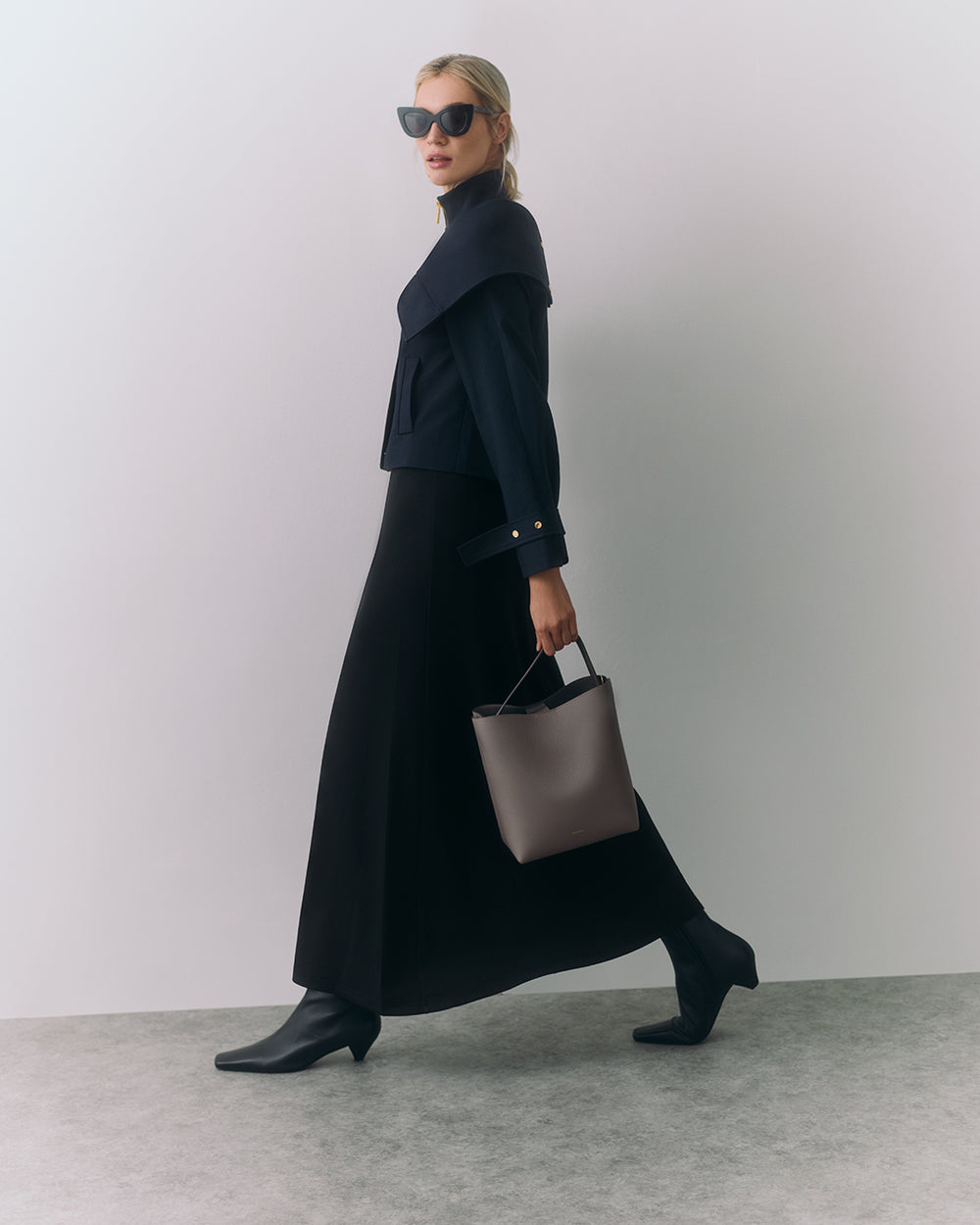 Woman in long skirt and jacket holds a handbag, wearing sunglasses and ankle boots.