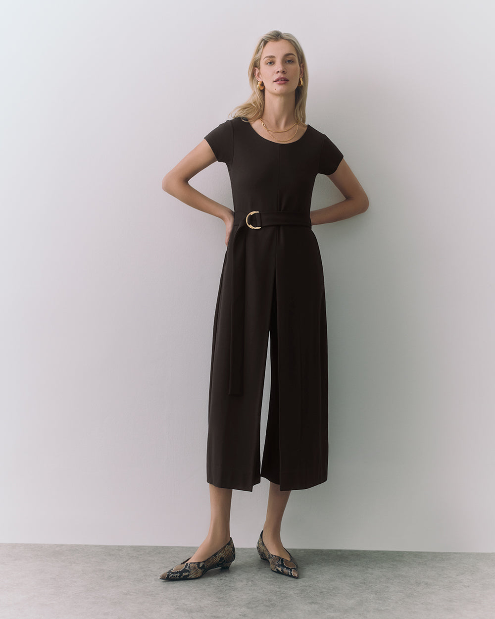 Person wearing a belted jumpsuit and flats stands against a wall.