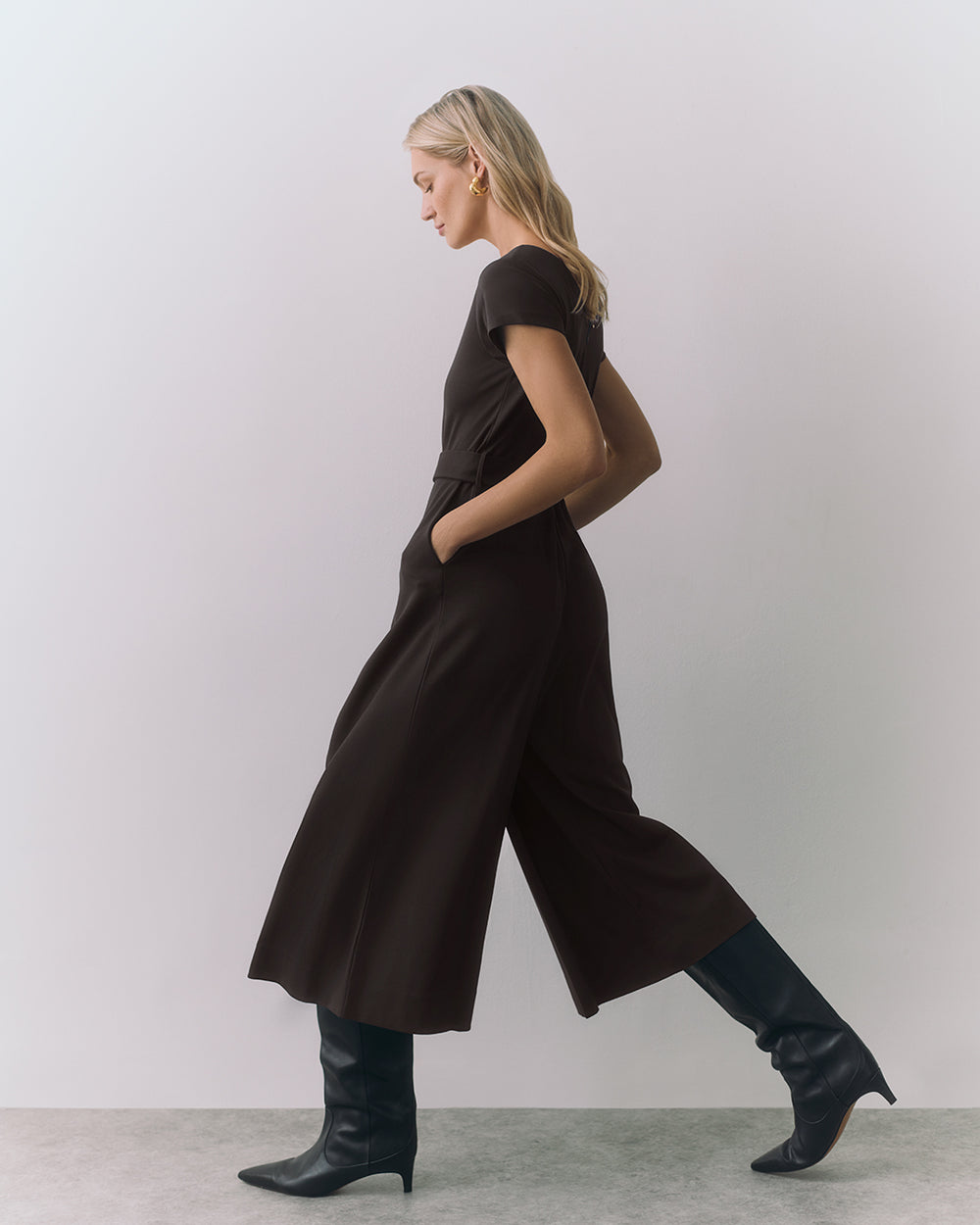 Person in a jumpsuit walking in boots against a plain background.