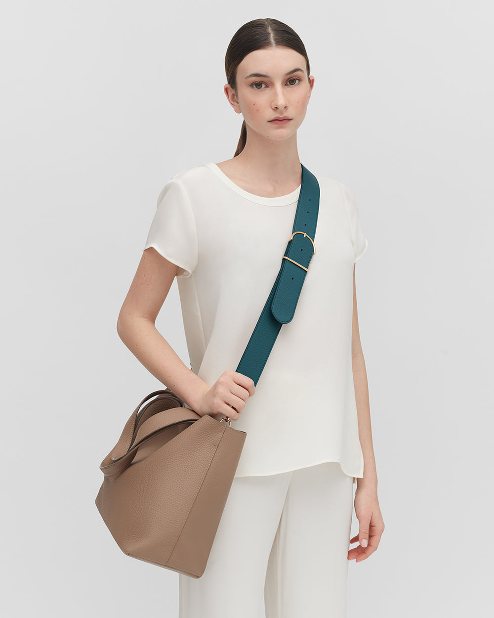 Woman in a plain top and pants holding a bag with a wide strap over her shoulder.