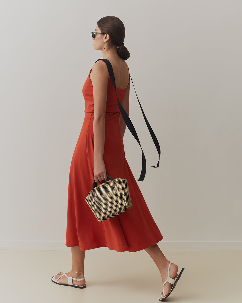 Woman walking in dress, wearing sunglasses and sandals, holding a woven handbag.