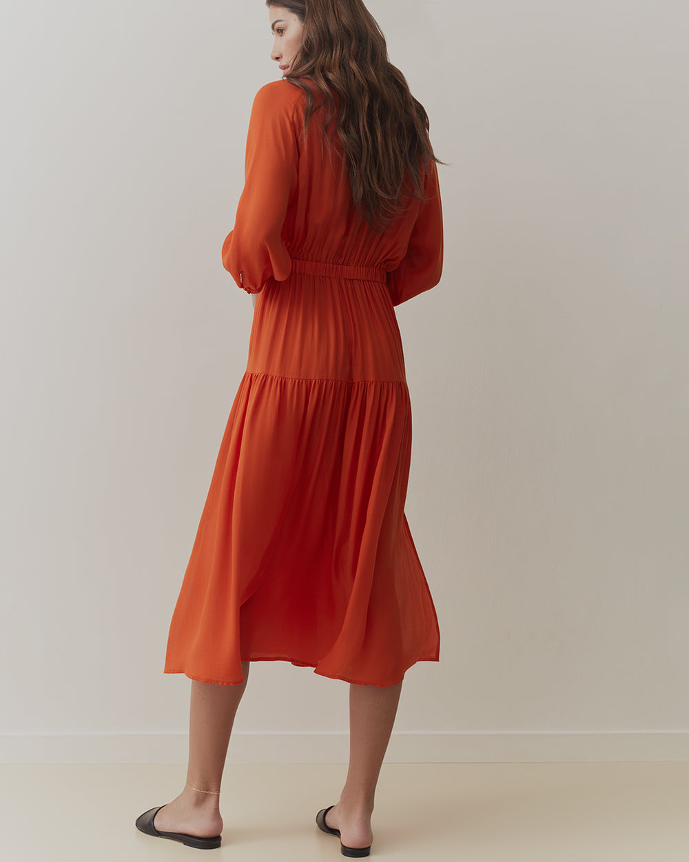Woman in a long-sleeved dress, viewed from the back, standing indoors.