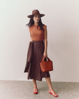 Woman in sleeveless top, long skirt, hat, and sandals holding a handbag.