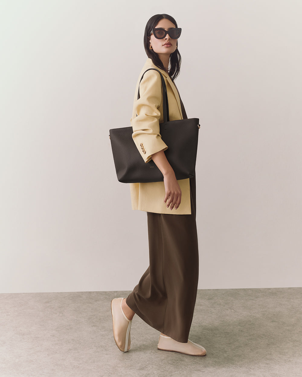 Person in a blazer with a large shoulder bag and sunglasses standing indoors