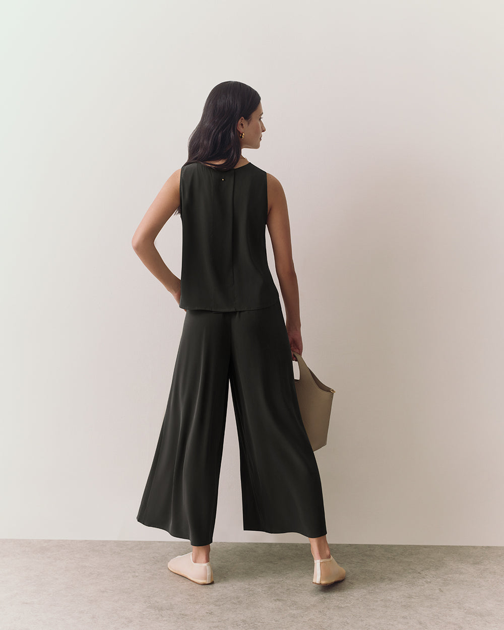 Person wearing sleeveless top and wide-leg pants holding a bag, standing on a plain floor.