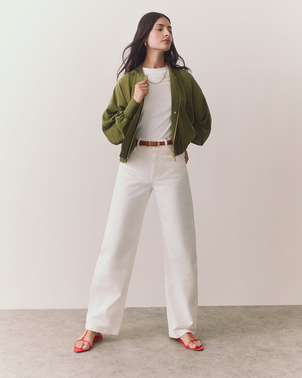 Person wearing open jacket, t-shirt, wide pants, sandals, and necklace standing confidently