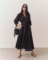 Woman in long dress with sunglasses holds a woven handbag and stands on a plain background.