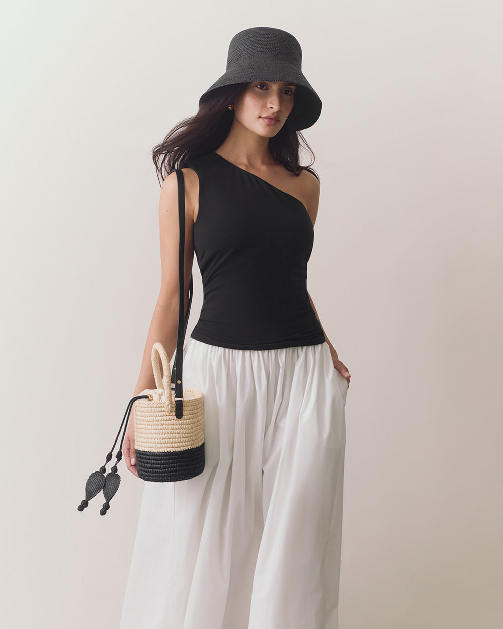Woman wearing a hat, one-shoulder top, wide pants, carrying a small woven bag.