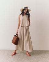 Woman in sleeveless top and wide pants holding a tote bag, wearing a hat and sandals.