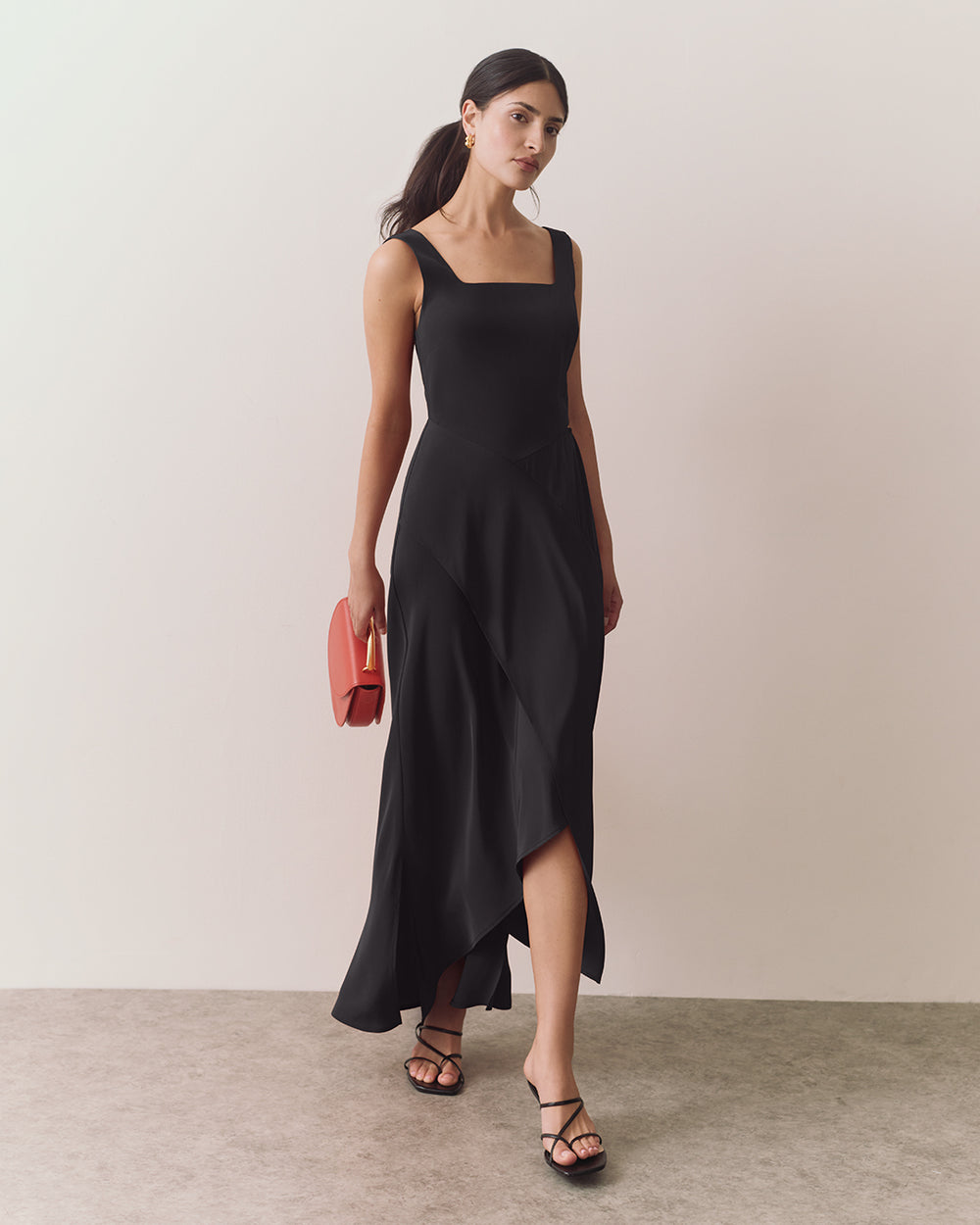 Woman in long dress holding handbag, wearing sandals, standing against plain background.