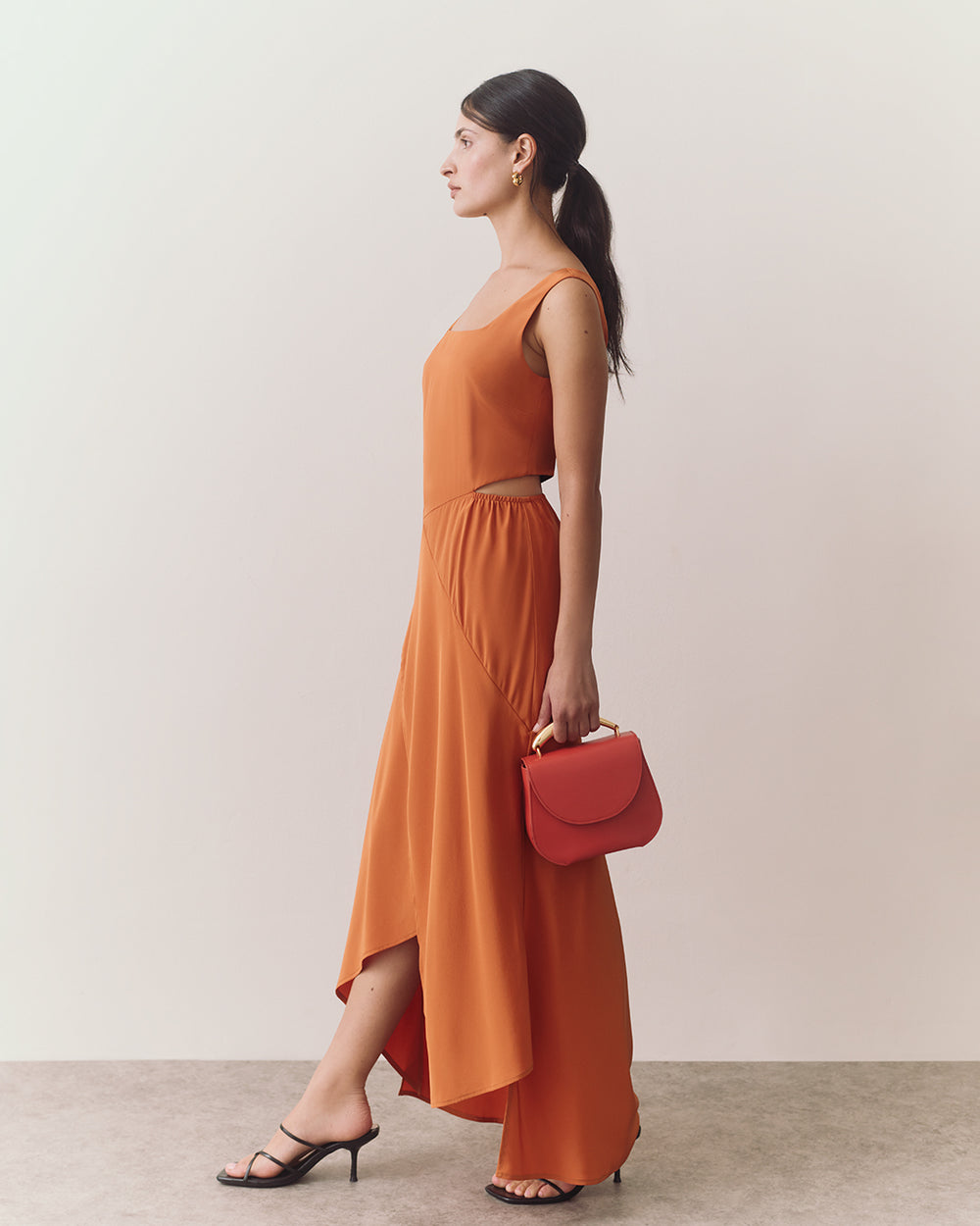 Woman in long dress holding small handbag, wearing heeled sandals, standing sideways.