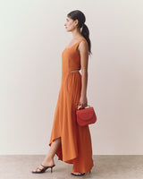 Woman in long dress holding small handbag, wearing heeled sandals, standing sideways.