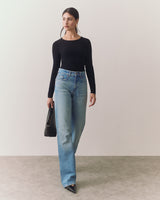 Person in long-sleeve top and jeans holding a handbag, standing indoors.