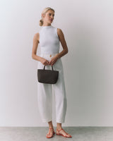 Person standing, holding a small bag, wearing a sleeveless top and pants, with sandals.