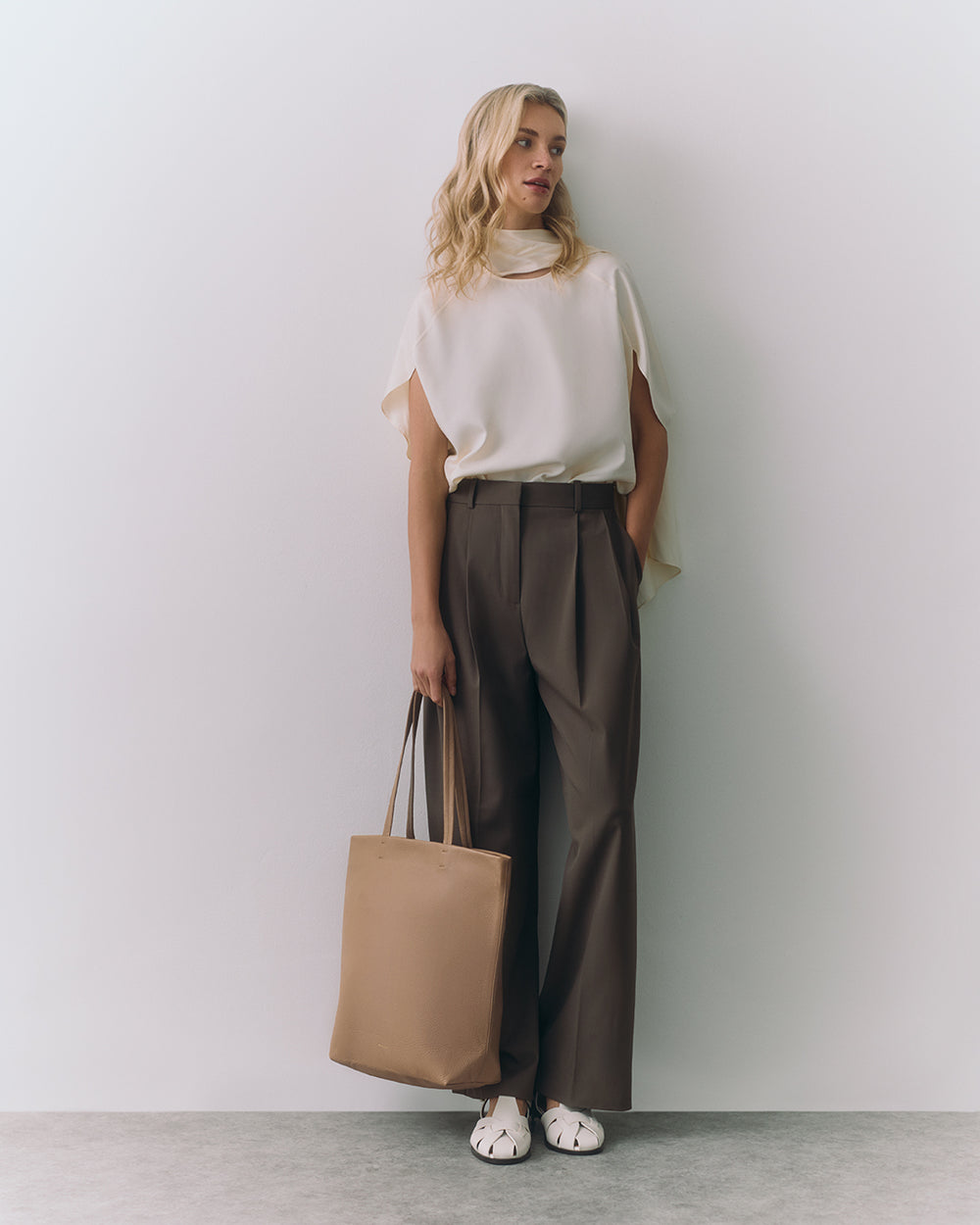 Person wearing a top and trousers holding a tote bag, standing against a plain backdrop.