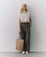 Person wearing a top and trousers holding a tote bag, standing against a plain backdrop.