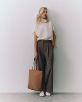 Person in a sleeveless top, trousers, holding a large tote bag, standing against a plain wall.
