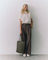 Woman standing against wall holding a tote bag and wearing a sleeveless top with wide-leg pants.