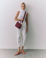 Person standing against wall holding a handbag over shoulder, wearing sleeveless top and pants.