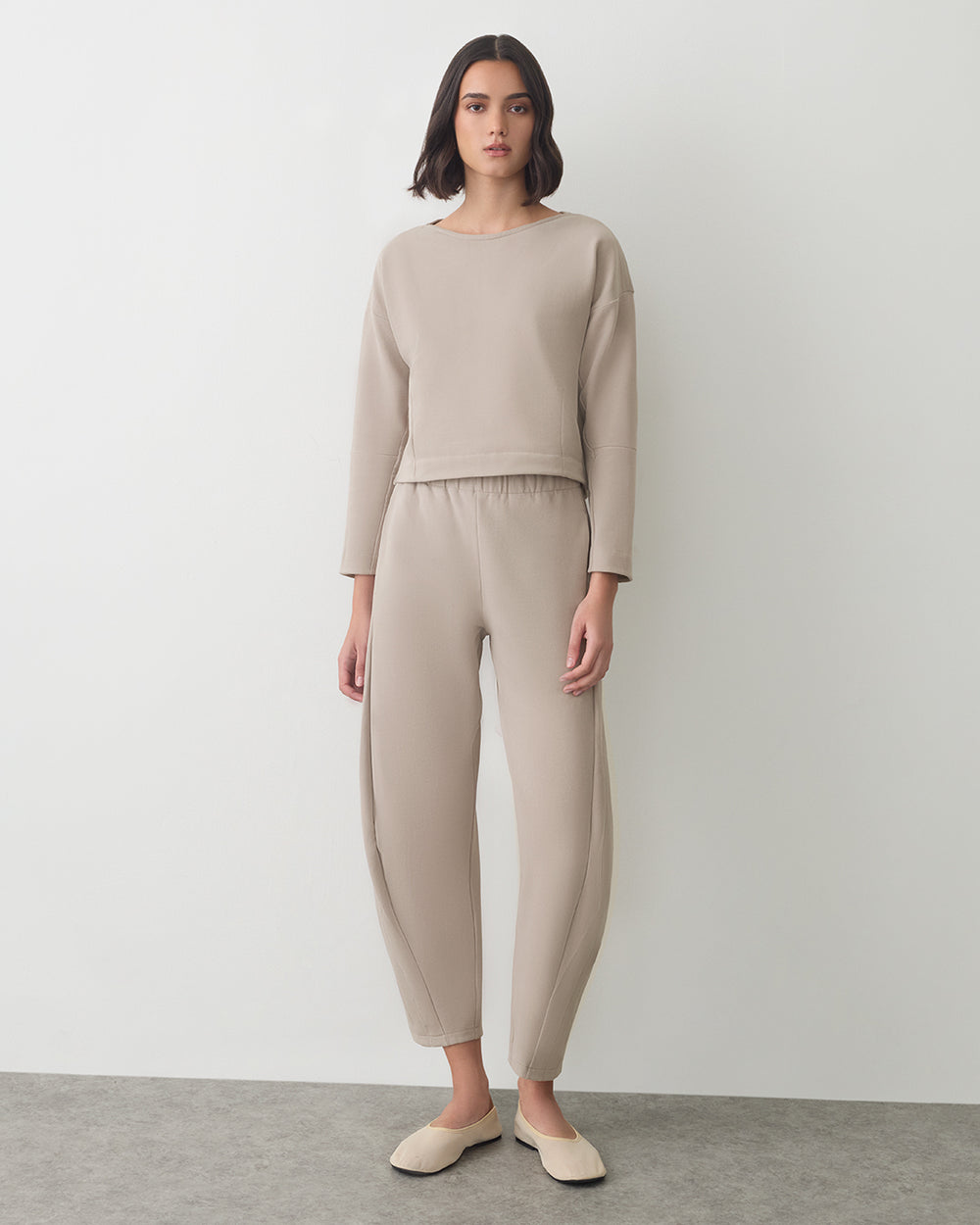 A person in a plain long-sleeve top and pants with slip-on shoes, standing in a neutral space.