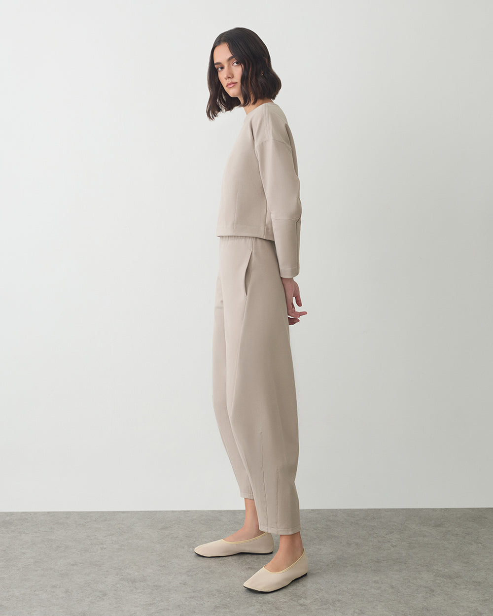 Person stands wearing a long-sleeve top and pants with flat shoes.