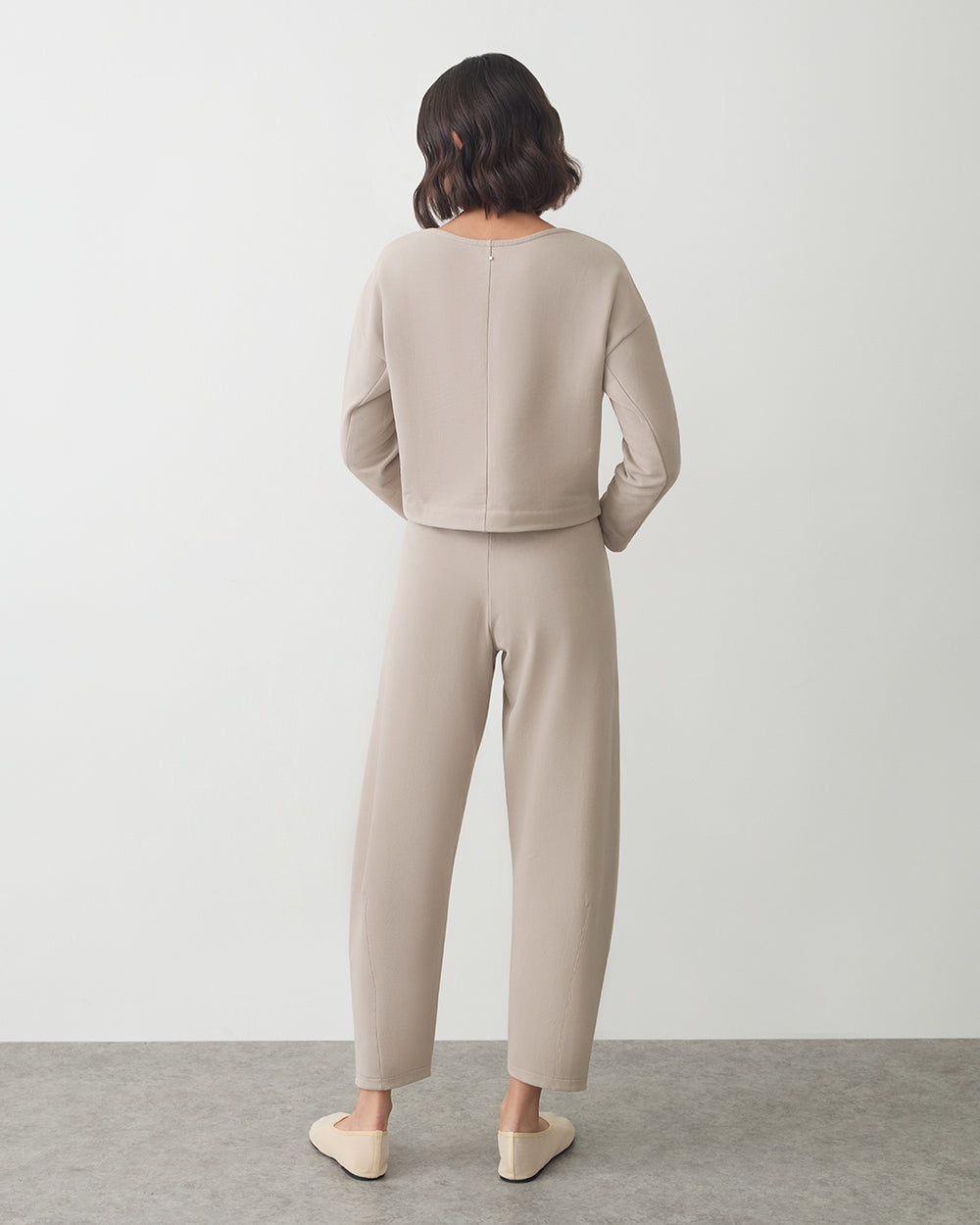 Person wearing long-sleeve top and pants stands on gray floor facing away.