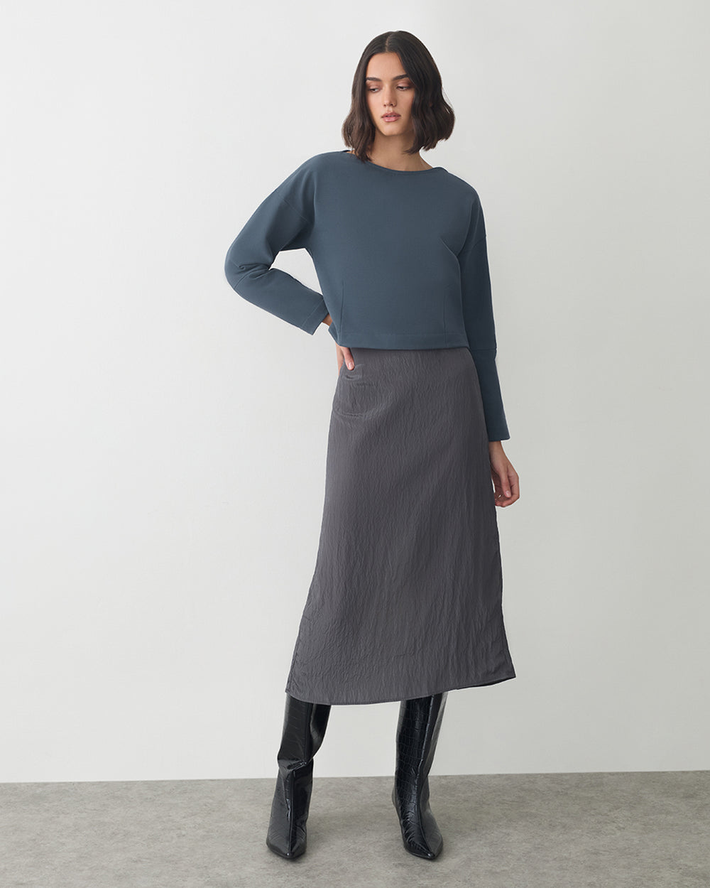 Woman in long skirt, boots, and long-sleeved top stands on carpeted floor.