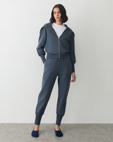 Person wearing a zip-up jacket, pants, and slip-on shoes stands against a plain backdrop.