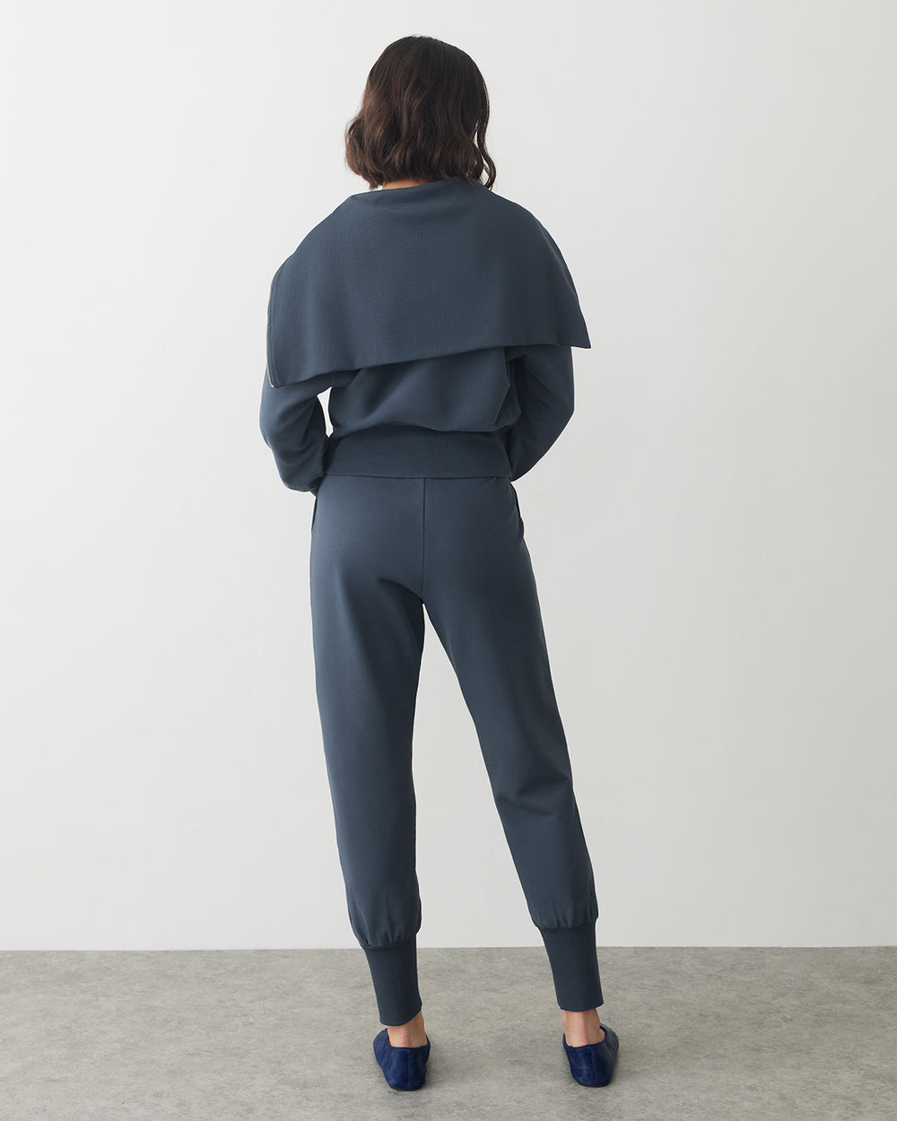 Person in matching outfit with cape, facing away, standing in a neutral space.