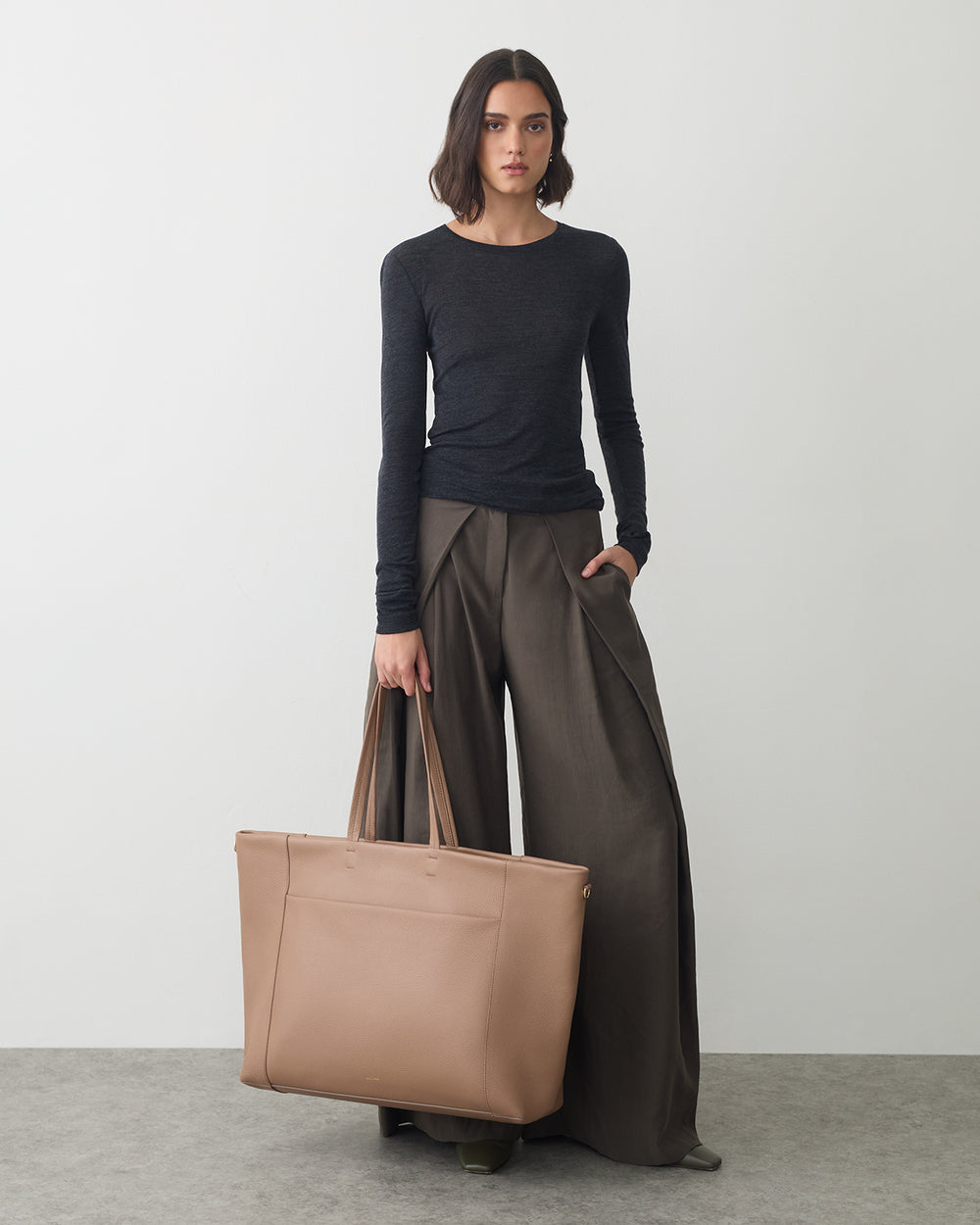 Person in long-sleeved top holds large travel bag and stands on gray floor.