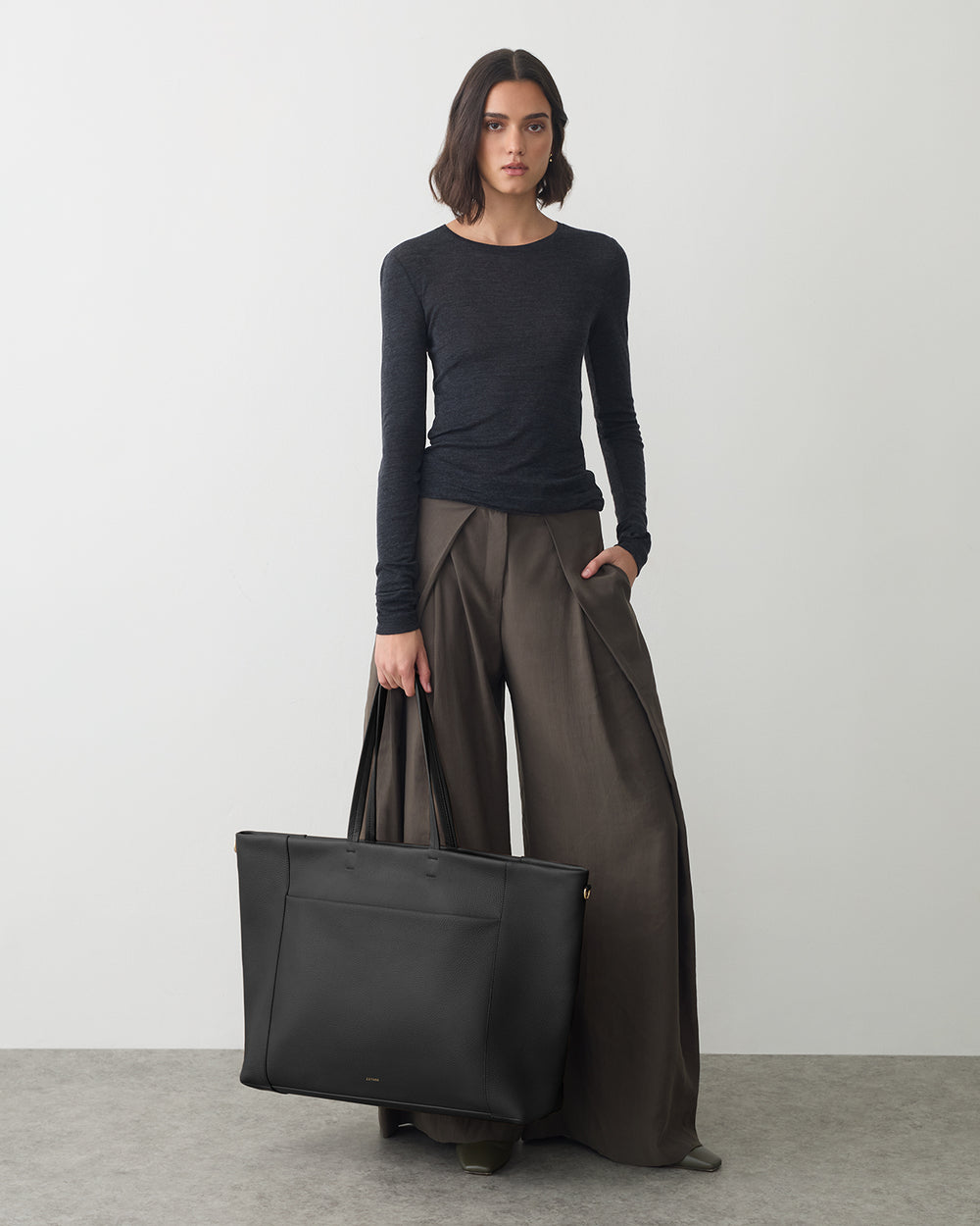 Person with long-sleeve shirt, wide pants, holding large bag standing indoors