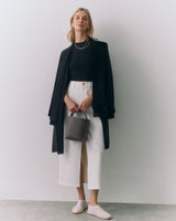 Person wearing a top, cape, pants, holding a small bag, standing against a plain backdrop.